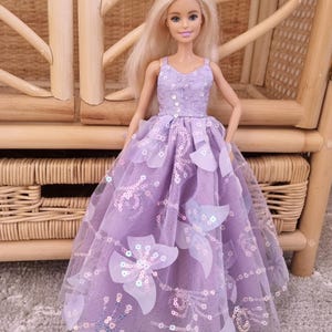 May include: A doll wearing a lavender dress with a floral design. The dress features a sequined bodice and a tulle skirt adorned with large, white flower appliqu&eacute;s. The doll has blonde hair and is posed in front of a wicker chair.