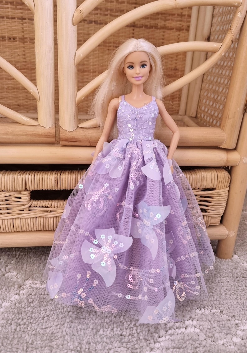 May include: A doll wearing a lavender dress with a floral design. The dress features a sequined bodice and a tulle skirt adorned with large, white flower appliqu&eacute;s. The doll has blonde hair and is posed in front of a wicker chair.