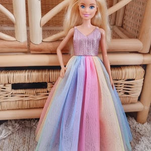 May include: A doll with blonde hair and blue eyes wearing a sparkly pink top and a rainbow-colored tulle skirt. The doll is standing in front of a woven wicker chair and a matching storage unit.