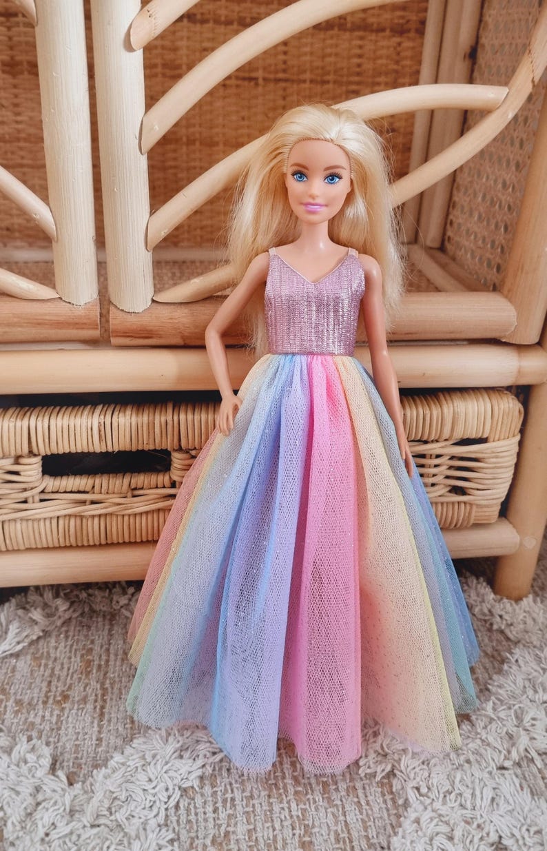 May include: A doll with blonde hair and blue eyes wearing a sparkly pink top and a rainbow-colored tulle skirt. The doll is standing in front of a woven wicker chair and a matching storage unit.