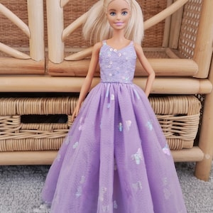May include: A doll wearing a lavender dress with a tulle overlay. The bodice is embellished with sequins, and the skirt features butterfly accents. The doll has blonde hair and is posed in front of a wicker chair.