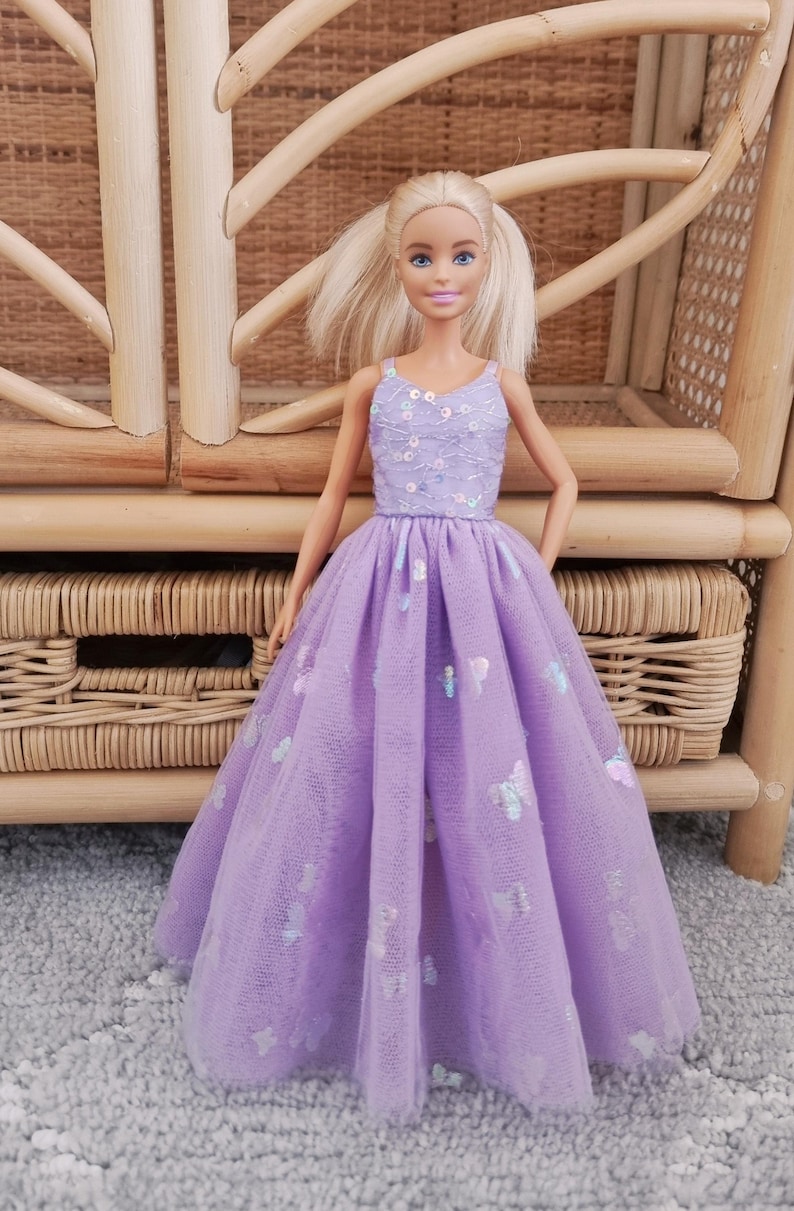 May include: A doll wearing a lavender dress with a tulle overlay. The bodice is embellished with sequins, and the skirt features butterfly accents. The doll has blonde hair and is posed in front of a wicker chair.
