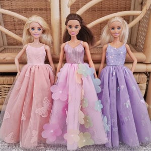 May include: Three dolls wearing pastel-colored gowns. The doll on the left is in a pink dress with butterfly accents. The middle doll wears a pink dress with flower accents. The doll on the right is in a lavender dress with sequin and butterfly accents.