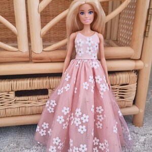 May include: A doll wearing a pink dress with white flower patterns. The dress has a tulle overlay and a fitted bodice. The doll has blonde hair and is standing in front of a wicker chair.