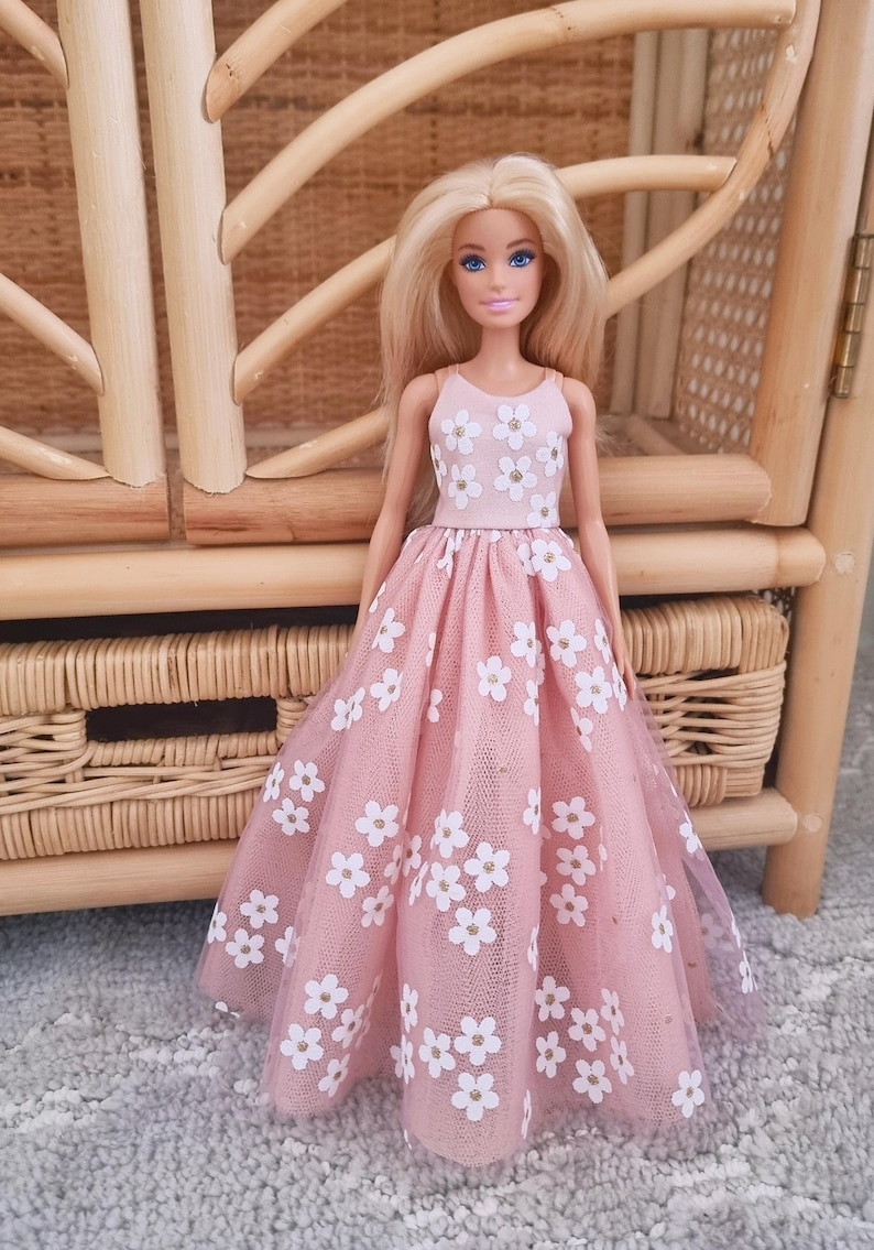 May include: A doll wearing a pink dress with white flower patterns. The dress has a tulle overlay and a fitted bodice. The doll has blonde hair and is standing in front of a wicker chair.
