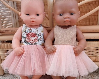 May include: Two baby dolls with distinct skin tones, each wearing a sleeveless dress with a full tulle skirt. The doll on the left wears a floral print bodice, while the doll on the right wears a shimmering gold bodice. Both skirts are light pink.