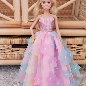 May include: A doll with blonde hair and blue eyes wearing a pink bodice and a long, flowing pink skirt adorned with pastel-colored flower appliqu&eacute;s. The doll is posed in front of a woven wicker chair.