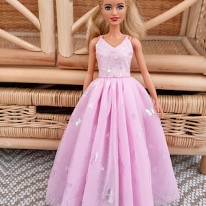May include: A doll wearing a pink dress with a V-neck bodice adorned with sequins and a flowing skirt embellished with silver butterfly accents. The doll has blonde hair and is posed in front of a woven wicker background.