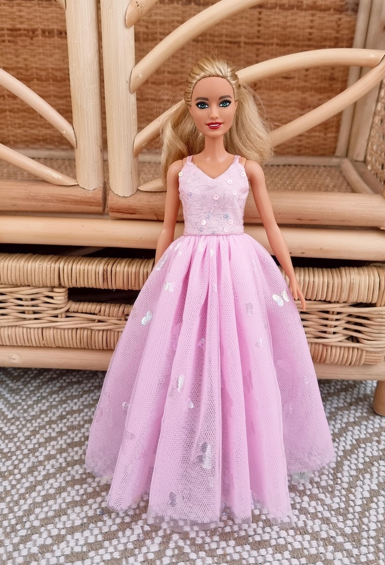 May include: A doll wearing a pink dress with a V-neck bodice adorned with sequins and a flowing skirt embellished with silver butterfly accents. The doll has blonde hair and is posed in front of a woven wicker background.