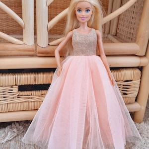 May include: A doll with blonde hair and blue eyes wearing a pink tulle gown with a gold bodice. The doll is posed in front of a wicker chair, creating a whimsical scene.