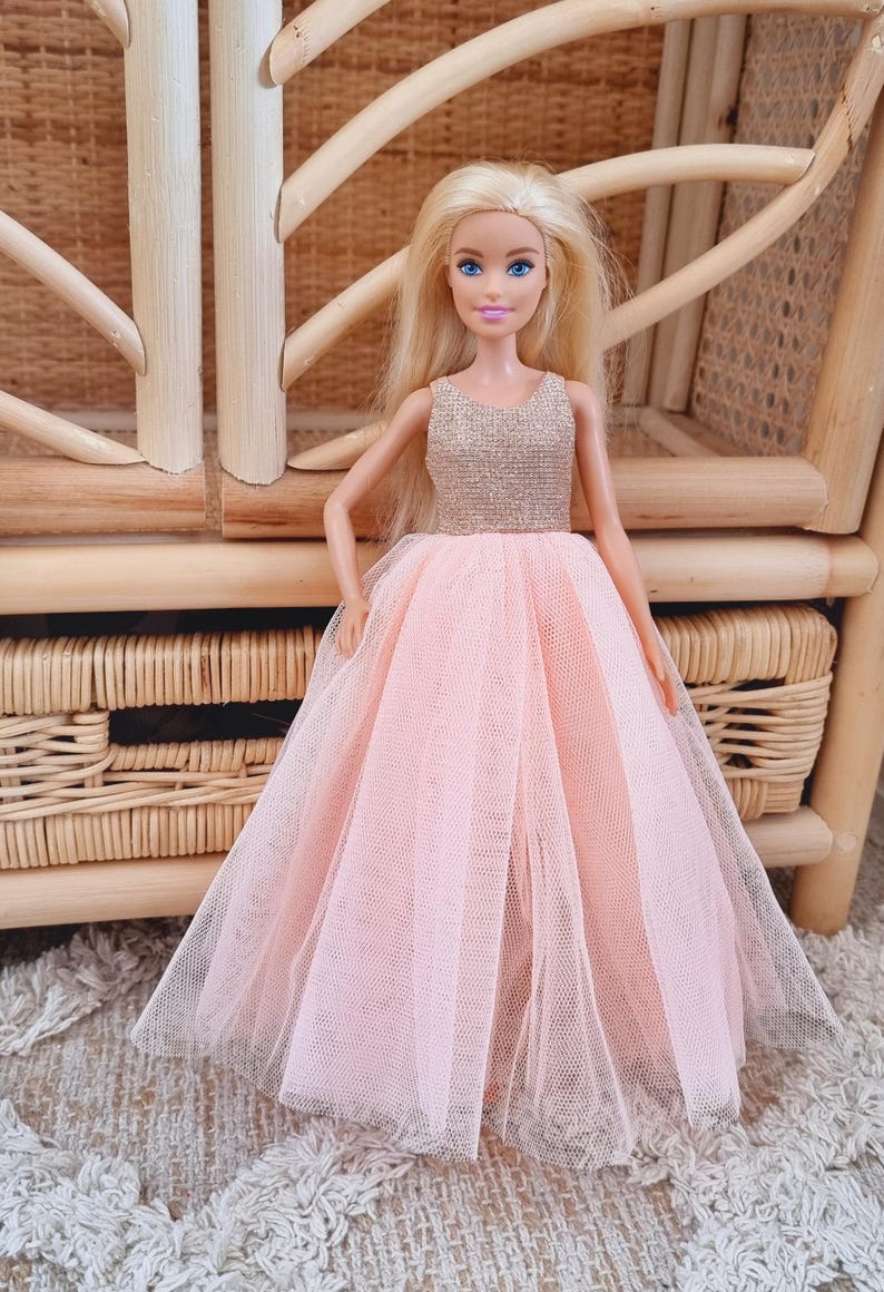 May include: A doll with blonde hair and blue eyes wearing a pink tulle gown with a gold bodice. The doll is posed in front of a wicker chair, creating a whimsical scene.