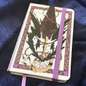 Puede incluir: Un diario blanco con una banda elástica morada y una cinta de marcador a juego. La portada presenta una ilustración en negro y dorado de una pluma, un reloj y un diseño de cadena con detalles morados y dorados. El diario está sobre una manta azul.