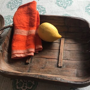 May include: A rustic, rectangular wooden tray with woven handles. Inside the tray are a folded orange linen napkin, and a yellow lemon. The tray is made of weathered wood and is suitable for serving food or displaying items.
