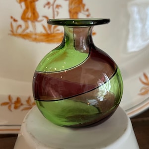 May include: A small, decorative glass vase with a rounded body and a narrow neck. The vase is divided into sections of green, brown, and burgundy, with a dark green rim. The vase is sitting on a white surface, with a decorative plate in the background.