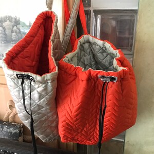May include: Two quilted tote bags, one orange and one silver, hanging from a wooden surface. Both bags have black drawstrings and straps.
