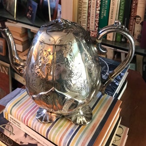 Op de afbeelding: Een zilveren theepot met een sierlijk ontwerp en een bolvormig lichaam. De theepot staat op een stapel kleurrijke gestreepte boeken.