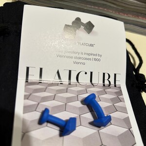 Może przedstawiać: Dwie niebieskie spinki do mankietów o geometrycznym wzorze są wyświetlane na karcie z napisem "FLATCUBE". Karta zawiera również tekst "Cufflinks 'FLATCUBE'" i "jewellery is inspired by Viennese staircases | 1900 Vienna". Tło to wzór szarych i białych kształtów geometrycznych.