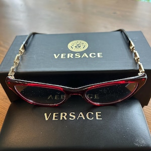 Może przedstawiać: Okulary w czerwonej oprawie ze złotymi akcentami na zausznikach, spoczywające na czarnym etui i pudełku. Na pudełku i etui widnieje nazwa marki "VERSACE" złotymi literami. Okulary mają kształt kociego oka.