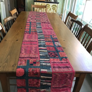 May include: A red and blue patterned table runner with tassels on a wooden table. The runner is made of fabric and has a patchwork design.