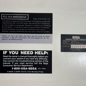 May include: A white sheet with three black rectangular stickers. The top sticker has the text "FCC ID: FJ8MD2SEGA". The middle sticker reads "IF YOU NEED HELP:". The bottom sticker has the text "SEGA GENESIS SYSTEM CONSOLE".