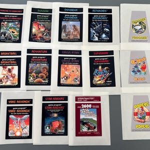 May include: A collection of 12 Atari 2600 game cartridge labels. The labels are printed on white paper with black text and graphics. The labels feature popular Atari games such as Pac-Man, Donkey Kong, and Space Invaders.
