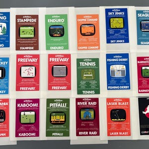 May include: A collection of 16 vintage Activision video game cartridges for the Atari 2600 console. The cartridges are arranged in four rows of four, with each cartridge featuring a different game title and artwork. The titles include Skiing, Stampede, Enduro, Chopper Command, Sky Jinks, Seaquest, Ice Hockey, Freeway, Tennis, Fishing Derby, Barnstorming, Megamania, Kaboom!, Pitfall!, River Raid, Laser Blast, and Ghostbusters.