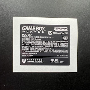 May include: A black and white Game Boy Player label with the Nintendo GameCube logo. The label includes the text "GAME BOY PLAYER" and other technical information, including "DOL-017" and "DOL-001".