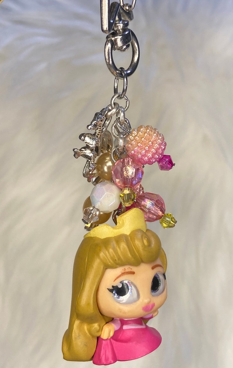 May include: A pink and yellow princess charm with a silver chain and beads. The charm features a cartoon princess with a crown, pink dress, and blonde hair.