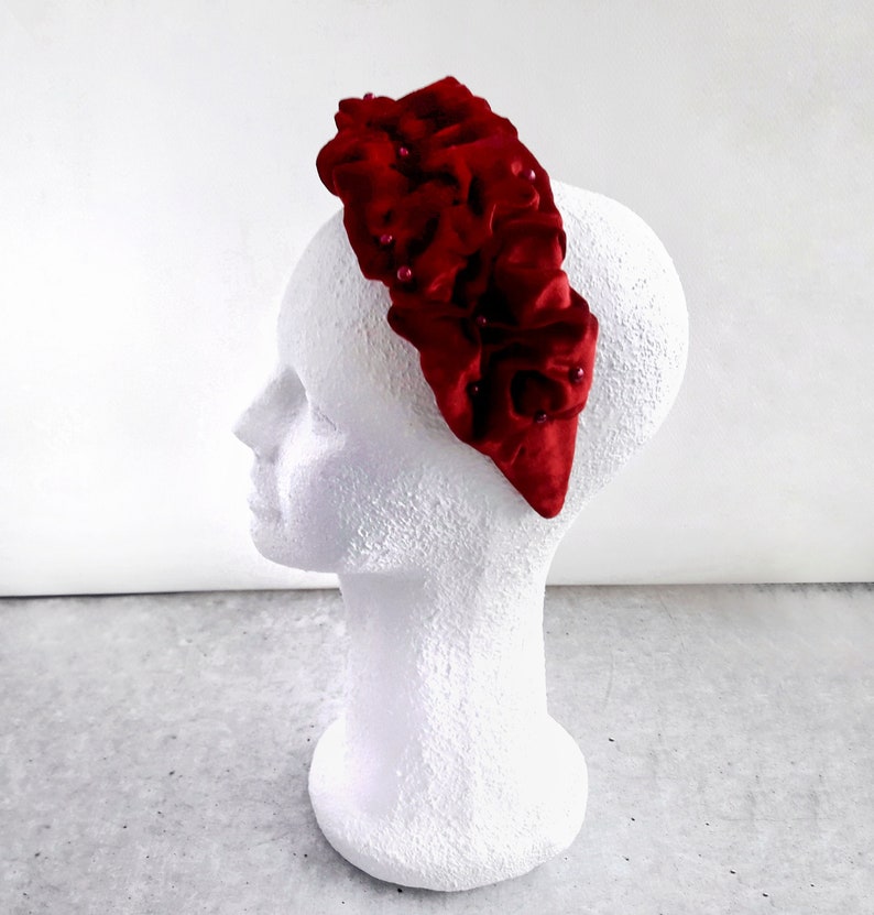 Puede incluir: Una diadema de terciopelo rojo con un dise&ntilde;o de volantes y peque&ntilde;os adornos de perlas.