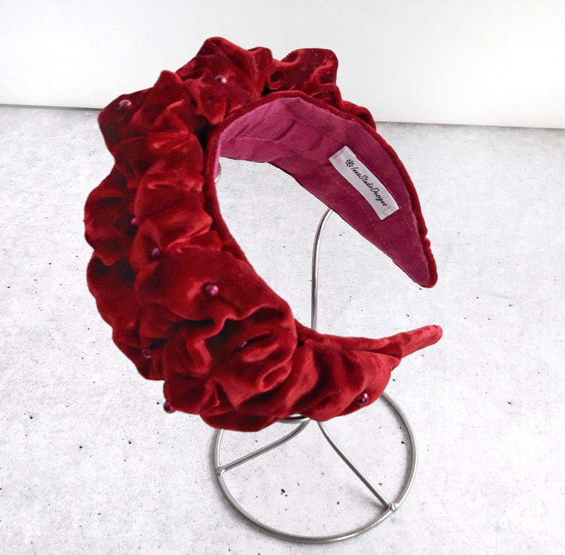 Puede incluir: Una diadema de terciopelo rojo con un dise&ntilde;o fruncido y peque&ntilde;os detalles de perlas. La diadema est&aacute; en un soporte de metal. La etiqueta en la diadema dice "#HandmadeDesign".