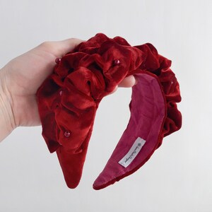 Puede incluir: Una diadema de terciopelo rojo con un dise&ntilde;o fruncido y peque&ntilde;as perlas. La diadema est&aacute; forrada con un tejido rosa y tiene una etiqueta que dice "Handmade by...".