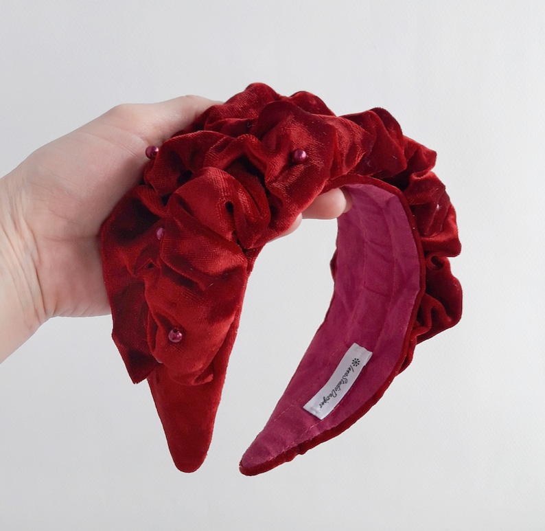 Puede incluir: Una diadema de terciopelo rojo con un dise&ntilde;o fruncido y peque&ntilde;as perlas. La diadema est&aacute; forrada con un tejido rosa y tiene una etiqueta que dice "Handmade by...".