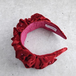 Puede incluir: Una diadema de terciopelo rojo con un dise&ntilde;o fruncido y peque&ntilde;os detalles de perlas. La diadema est&aacute; forrada con un tejido rosa y tiene una etiqueta que dice "The Crown Boutique Designer".