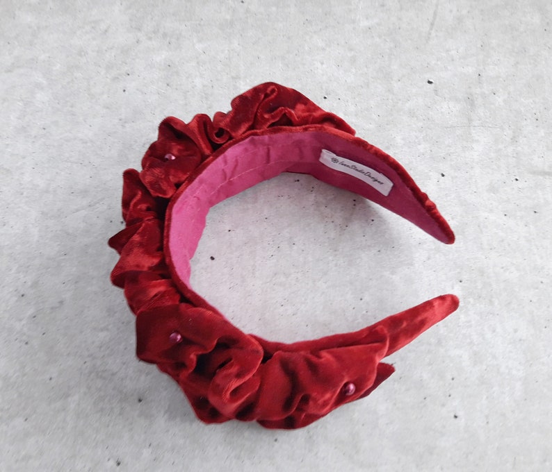 Puede incluir: Una diadema de terciopelo rojo con un dise&ntilde;o fruncido y peque&ntilde;os detalles de perlas. La diadema est&aacute; forrada con un tejido rosa y tiene una etiqueta que dice "The Crown Boutique Designer".