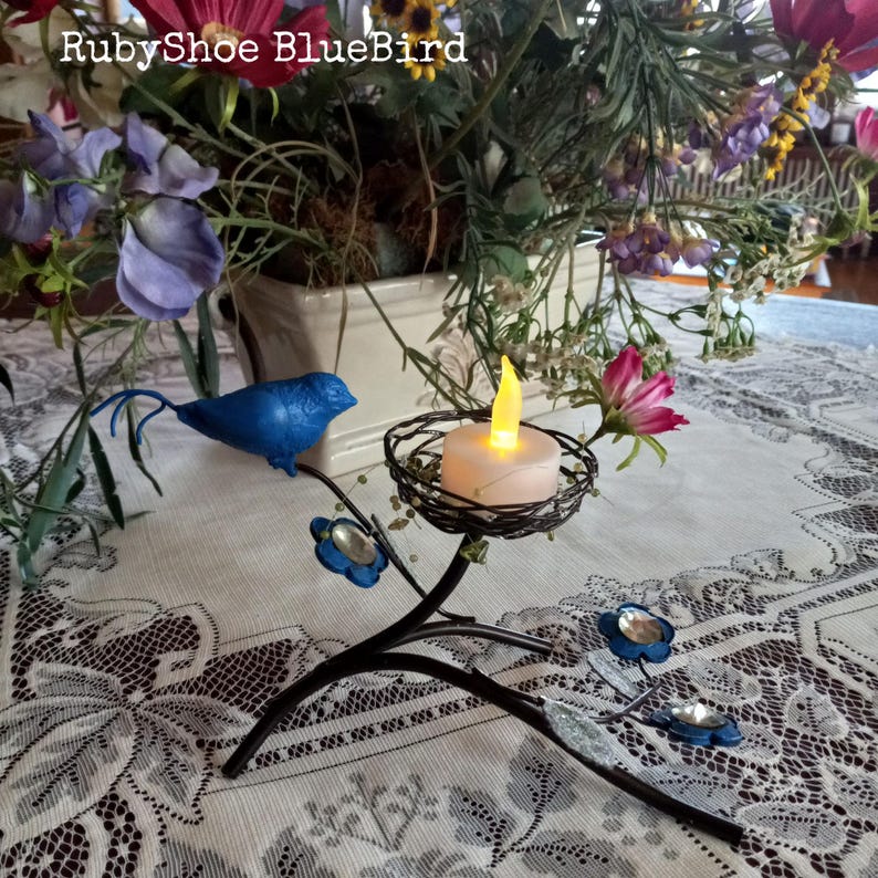 Może przedstawiać: Dekoracyjny metalowy świecznik z motywem ptaka i kwiat&oacute;w. Świecznik ma czarną metalową gałązkę z niebieskimi kwiatowymi akcentami i małym ptakiem. Świeca na baterie znajduje się w plecionym gnieździe.