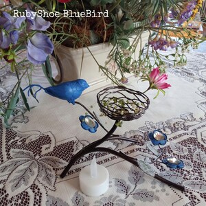Może przedstawiać: Dekoracyjna metalowa gałąź z niebieskim ptakiem, gniazdem i niebieskimi akcentami kwiatowymi. Gałąź trzyma białą świecę LED. Tekst "RubyShoe BlueBird" jest widoczny na zdjęciu.