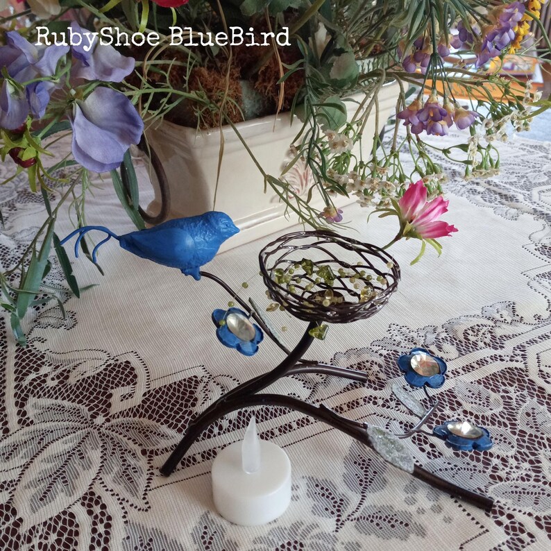 Może przedstawiać: Dekoracyjna metalowa gałąź z niebieskim ptakiem, gniazdem i niebieskimi akcentami kwiatowymi. Gałąź trzyma białą świecę LED. Tekst "RubyShoe BlueBird" jest widoczny na zdjęciu.