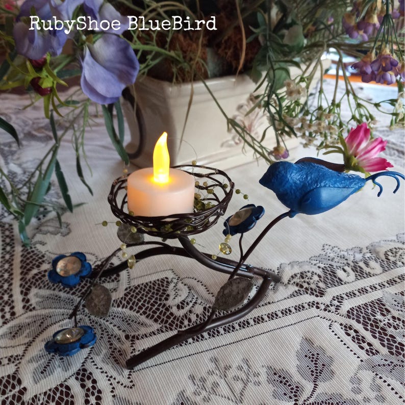 Może przedstawiać: Dekoracyjny świecznik z motywem niebieskiego ptaka, zasilany bateriami świecą w plecionym gnieździe. Uchwyt ma niebieskie kwiatowe akcenty i jest wykonany z metalu. Widoczny jest tekst "RubyShoe BlueBird".