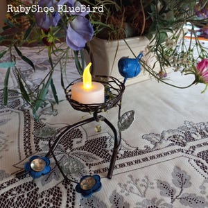 Może przedstawiać: Dekoracyjny świecznik z motywem ptaka i kwiat&oacute;w. Metalowy uchwyt posiada małą, okrągłą świecę z migoczącym ż&oacute;łtym światłem. Obecne są r&oacute;wnież niebieskie kwiatowe akcenty i niebieski ptak. Tekst "RubyShoe BlueBird" znajduje się na g&oacute;rze.