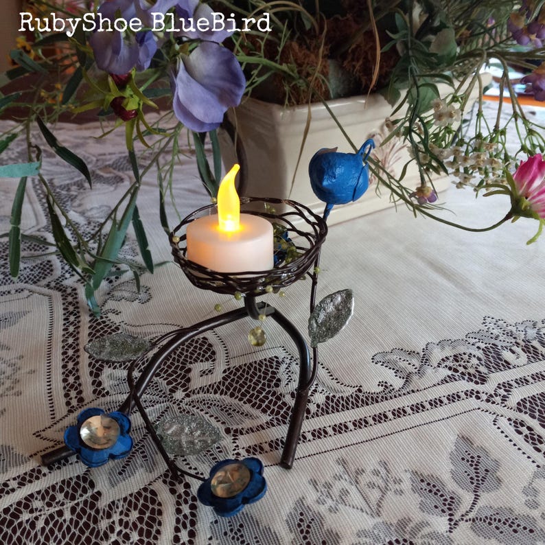 Może przedstawiać: Dekoracyjny świecznik z motywem ptaka i kwiat&oacute;w. Metalowy uchwyt posiada małą, okrągłą świecę z migoczącym ż&oacute;łtym światłem. Obecne są r&oacute;wnież niebieskie kwiatowe akcenty i niebieski ptak. Tekst "RubyShoe BlueBird" znajduje się na g&oacute;rze.