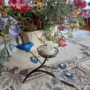 Może przedstawiać: Dekoracyjny metalowy świecznik z niebieskim ptakiem siedzącym na gałęzi, ozdobiony niebieskimi kwiatami. Zawiera białą świecę w małym, plecionym koszyku. Tekst "RubyShoe BlueBird" jest widoczny.