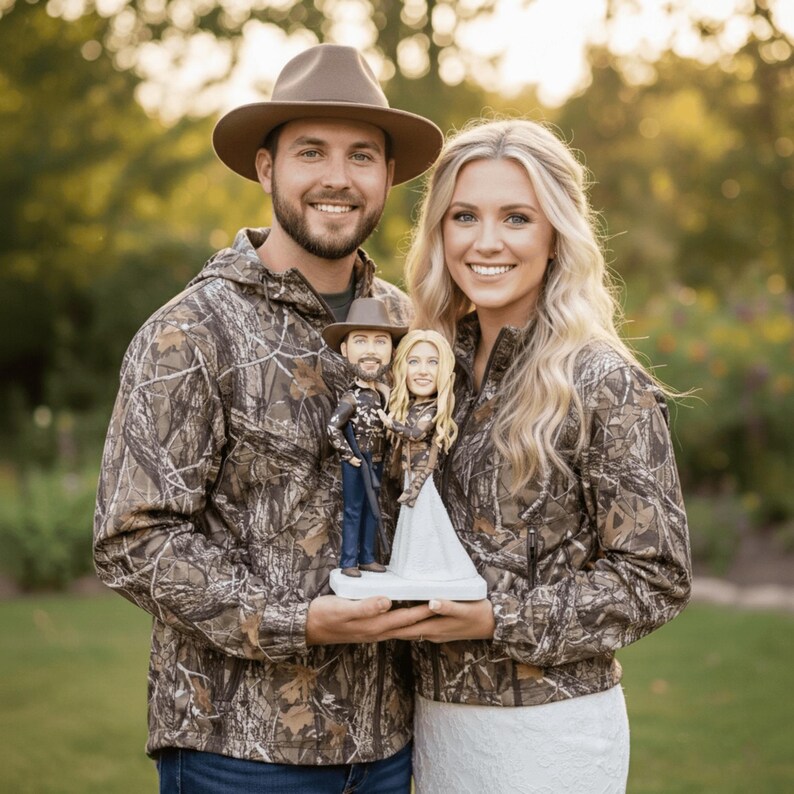 lifestyle photo showing the personalized hunter couple bobblehead being held by a real-life couple dressed in matching camouflage jackets, with an outdoor, sun-drenched background, perfect for a hunting themed wedding.