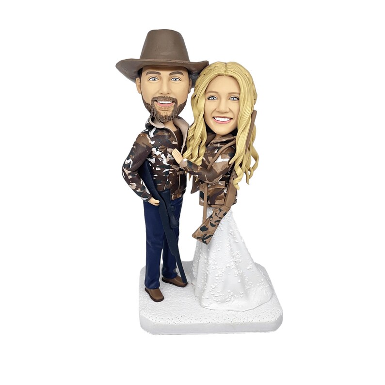 personalized hunter couple bobblehead cake topper on a white background, featuring a man in a brown hat and camouflage jacket holding a rifle, and a woman in a matching jacket and a white wedding dress.