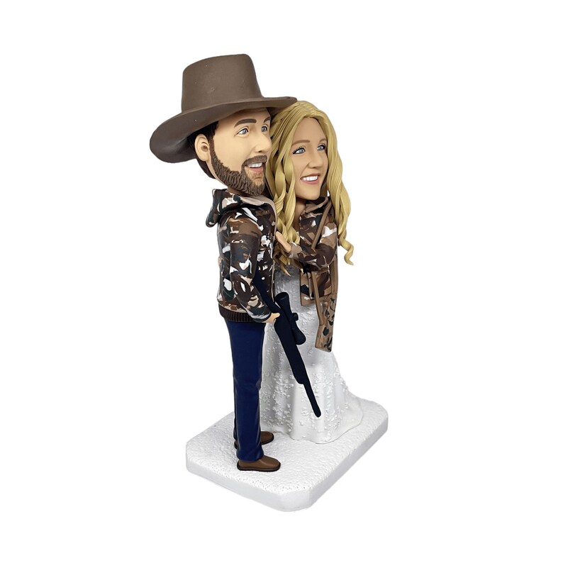 close-up side view of the custom bobblehead figurine, highlighting the facial details of the man and woman, their camouflage outfits, and the rifle, set against a plain white background.