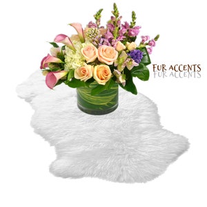 May include: A white faux fur rug with a bouquet of pink, white, and purple flowers in a clear glass vase. The vase is sitting on the rug. The text "FUR ACCENTS FUR ACCENTS" is in the upper right corner of the image.