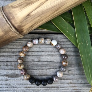 May include: A bracelet made of brown, white and grey stones with black lava beads. The bracelet is on a wooden surface with green leaves.