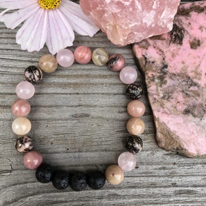 May include: A pink and black gemstone bracelet with lava beads. The bracelet is made with various shades of pink and black stones, including rose quartz and black lava beads. The bracelet is laid out on a wooden surface with pink and black gemstones.