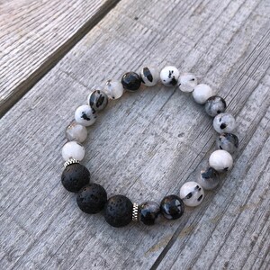 May include: A bracelet made of black lava rock and speckled white and black gemstone beads. The bracelet has silver-coloured metal accents and is strung on elastic. The beads are round and of varying sizes.