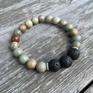 May include: A bracelet made of green, brown, and red stones with three black lava rock beads. The bracelet is on a wooden surface.