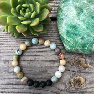 May include: A beaded bracelet featuring a mix of green, brown and white stones. The bracelet has a row of black lava beads at the bottom.